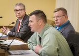 Justin Rost sits at the testifiers' table and speaks into a microphone to someone out of view. Gary Thaden and Representative Sundin sit next to him and watch him speak.